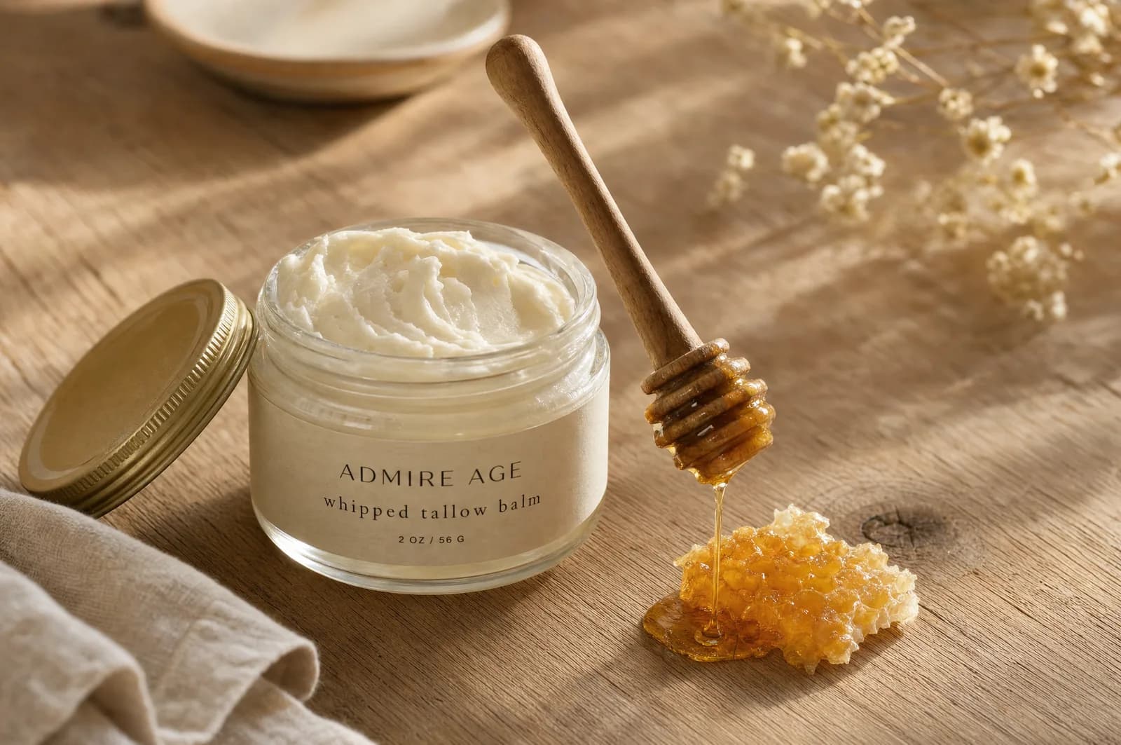 A jar of whipped tallow balm beside a wooden honey dipper drizzling golden raw honey, with a piece of honeycomb.