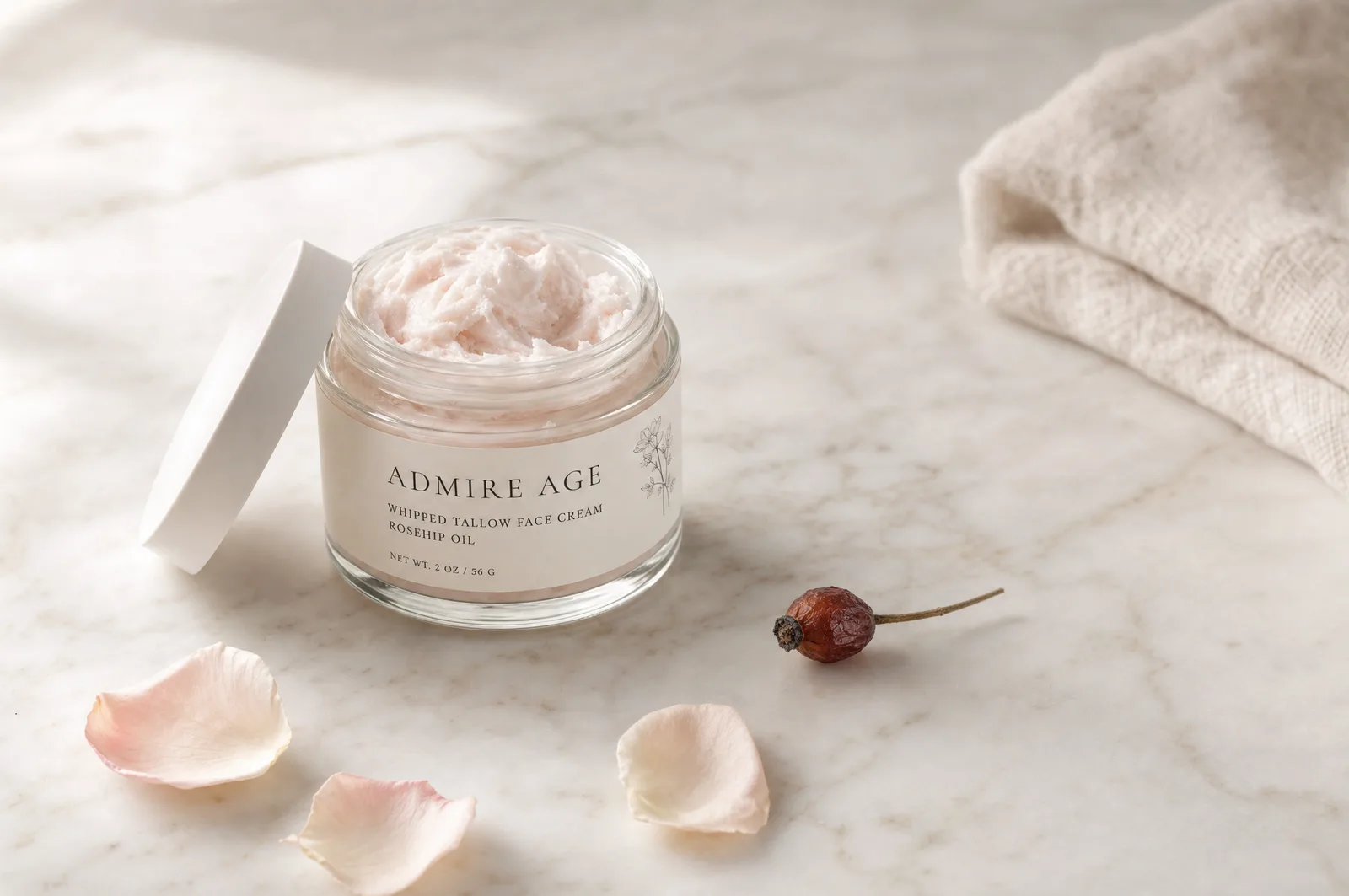 A small jar of airy, rose-tinted whipped tallow face cream with rose petals and a rosehip on pale marble.