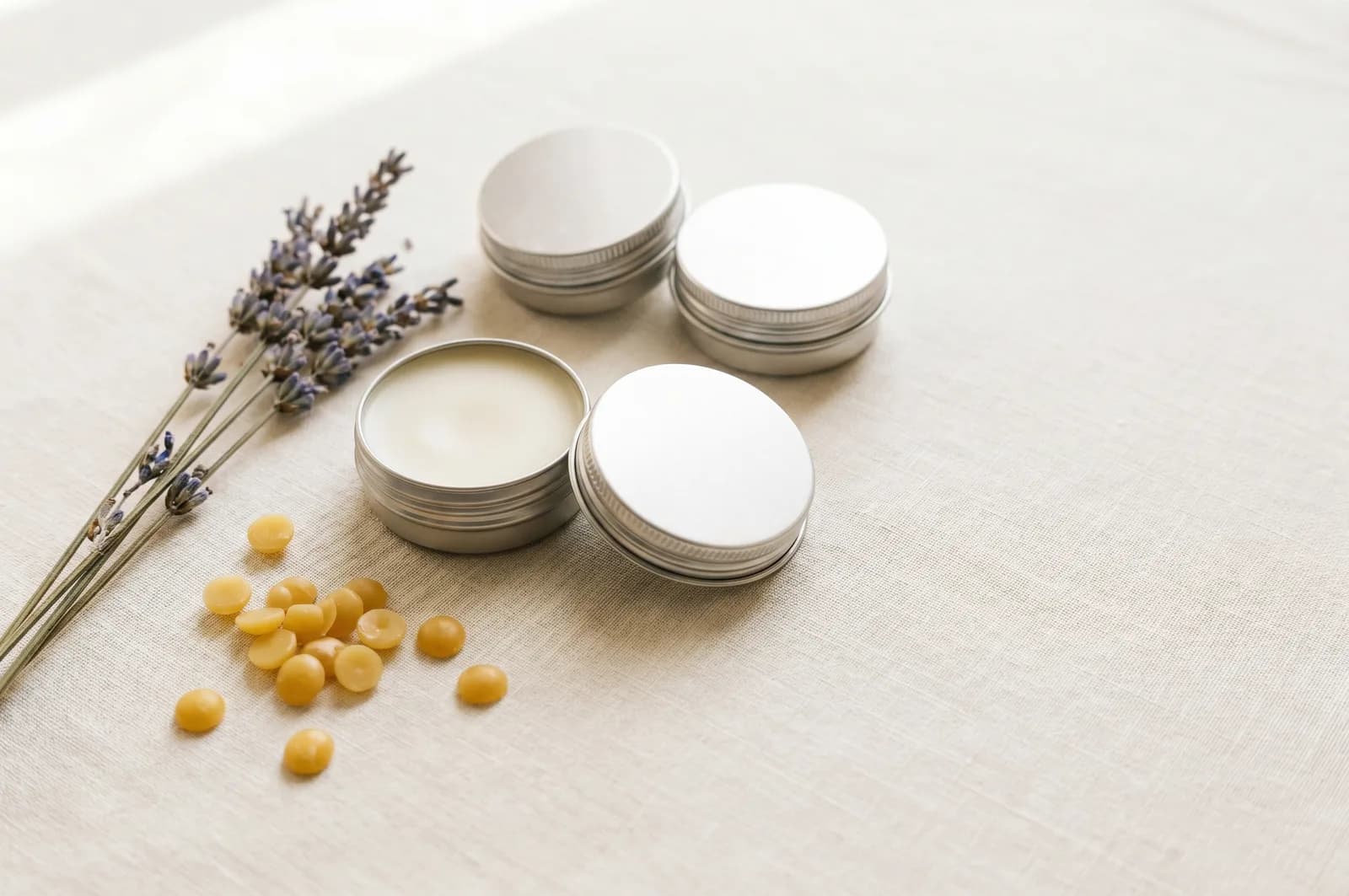 Small tins of homemade tallow lip balm on a pale linen surface beside a sprig of dried herbs.