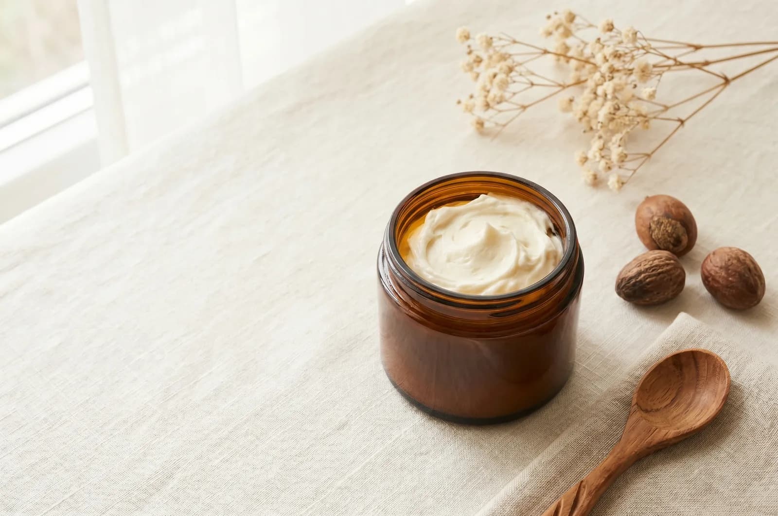 An open jar of softly whipped tallow body butter with soft peaks, on pale linen beside a few shea nuts.