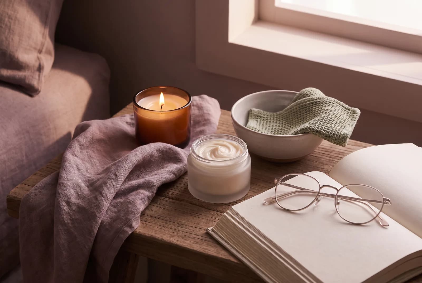 A lit candle, linen cloth, and open jar of tallow cream on a bedside table in soft evening light