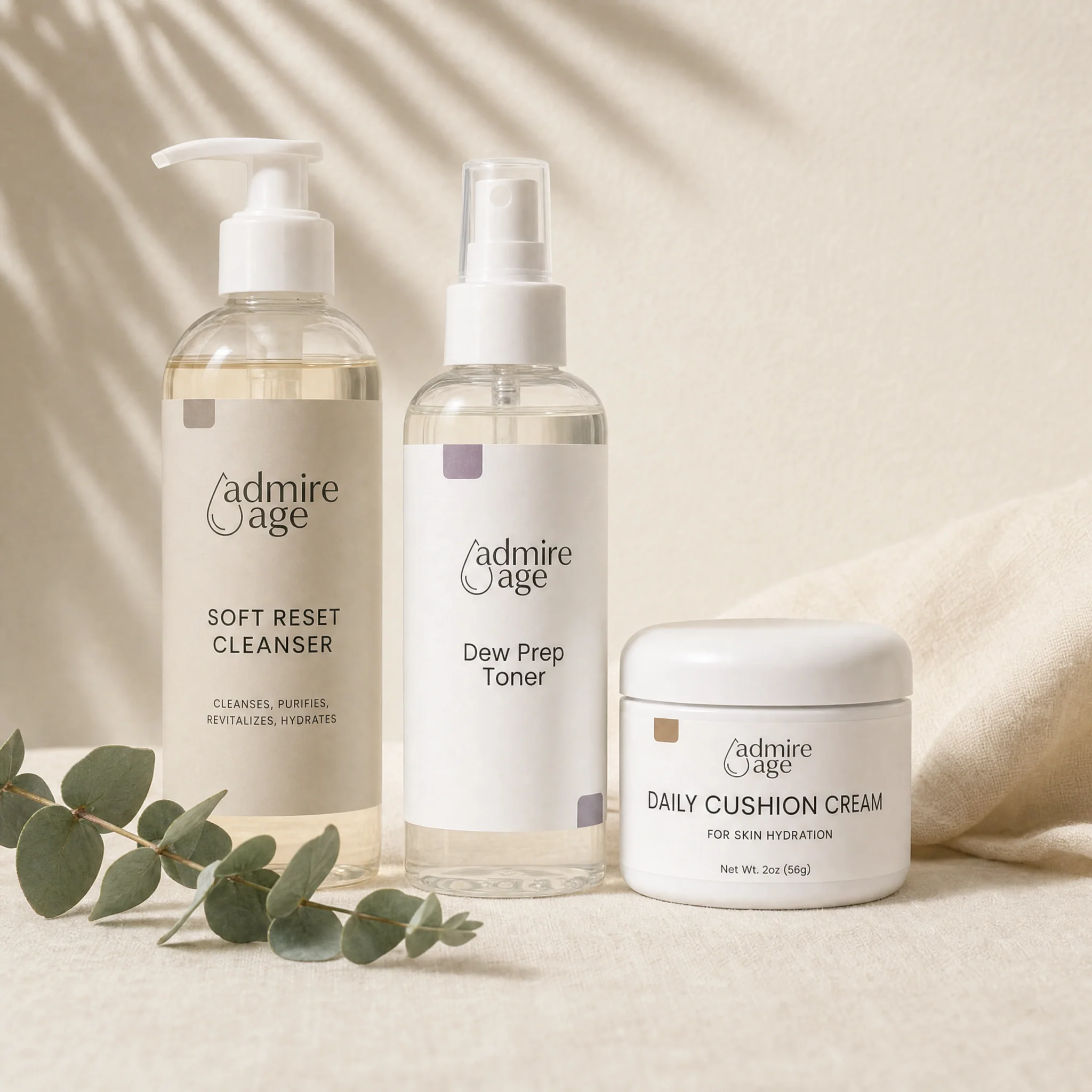 Admire Age Soft Reset Cleanser, Dew Prep Toner, and Skin Hydration Cream grouped together on a cream linen surface with a eucalyptus sprig