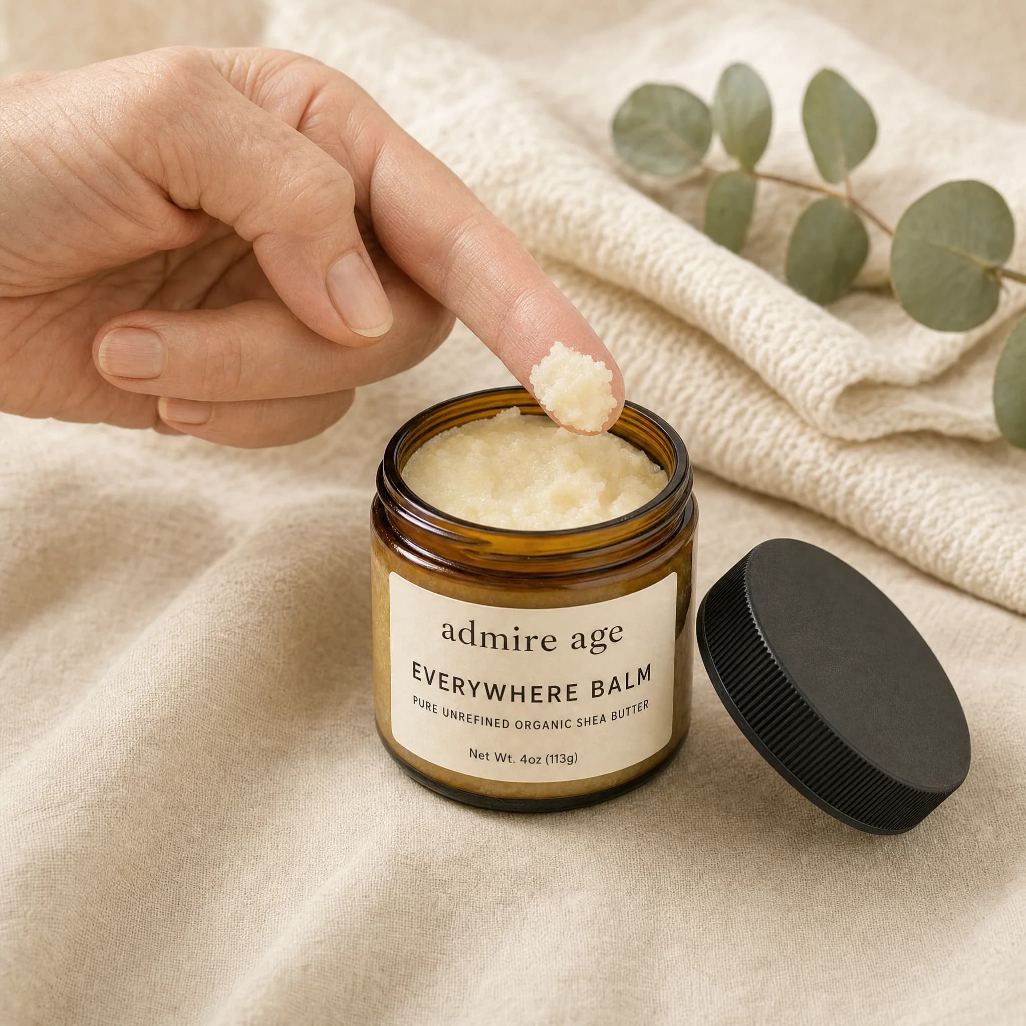 Mature woman's fingertip lifting a small scoop of off-white shea butter from the open Everywhere Balm amber glass jar on cream linen, knit towel and eucalyptus in background