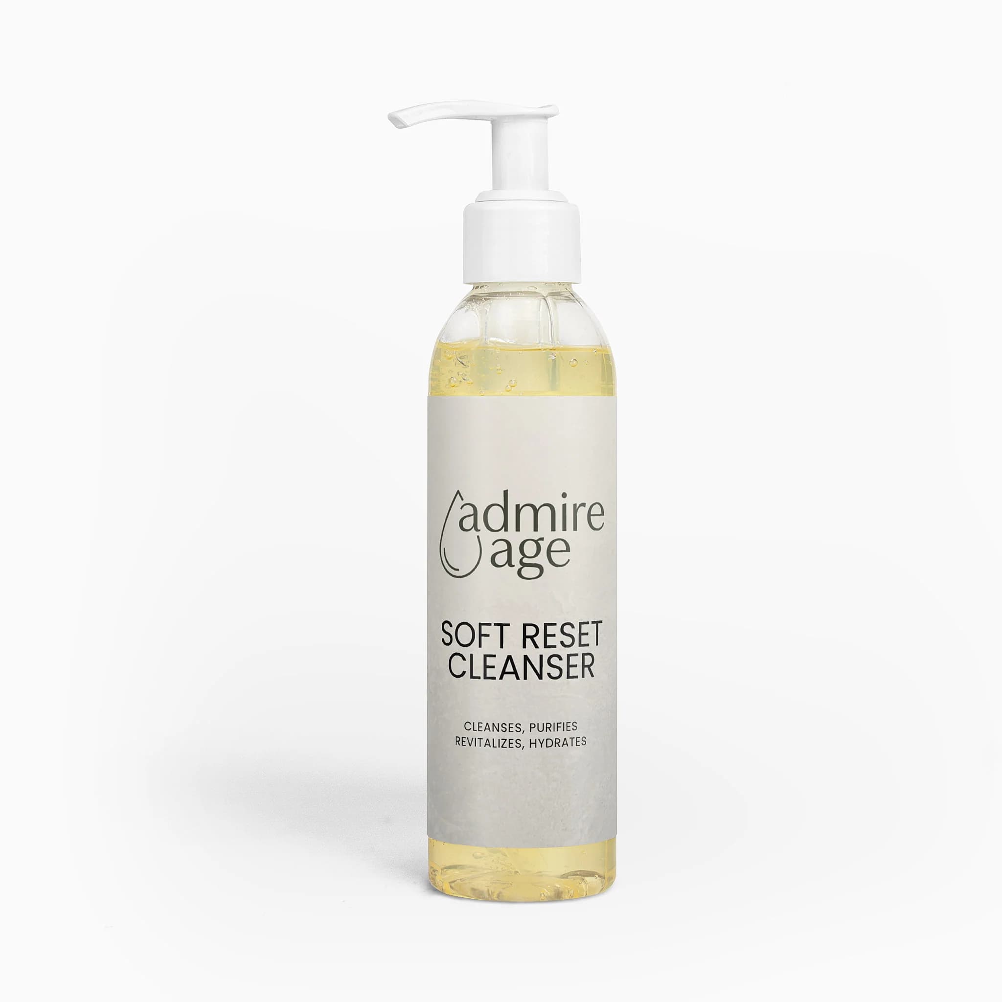 Front-on studio shot of the Admire Age Soft Reset Cleanser tall clear-glass pump bottle with a white pump cap and an ivory kraft-paper label reading admire age, SOFT RESET CLEANSER, cleanses purifies revitalizes hydrates, pale-gold gel visible inside