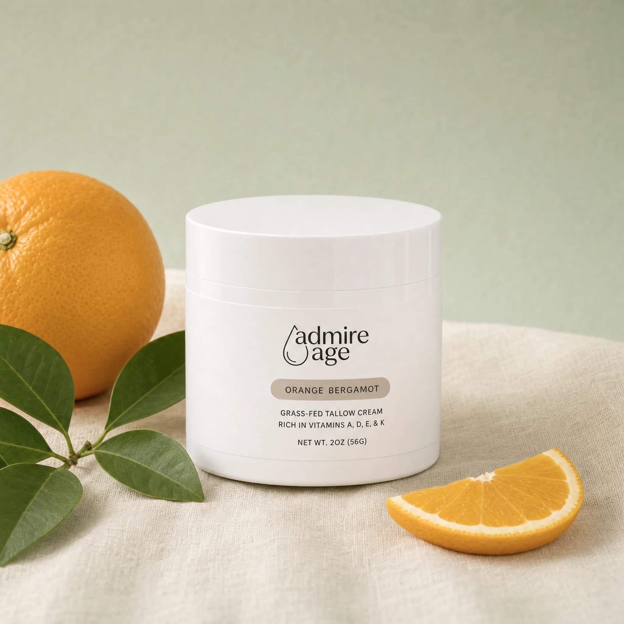 Admire Age Orange Bergamot tallow cream jar on a soft sage-green background with a fresh orange and citrus leaves