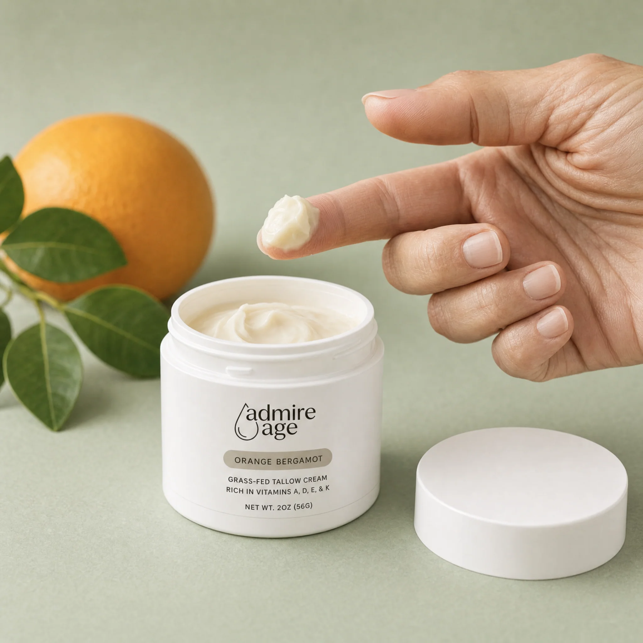 A hand lifting a small dollop of Orange Bergamot tallow cream from the open jar beside a fresh orange